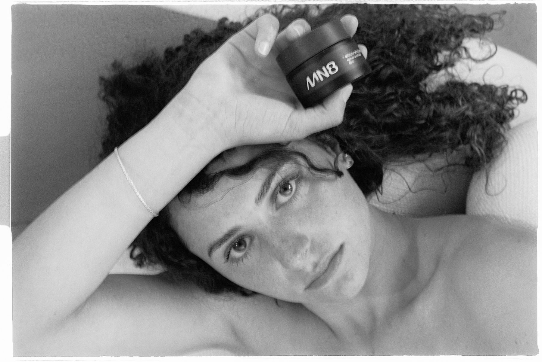 Woman lying down holding an MN8 Skincare jar, black and white